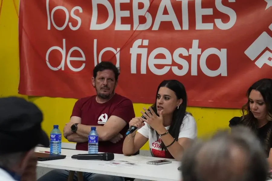 Tres personas sentadas en una mesa con micrófonos y botellas de agua participan en una mesa redonda. Detrás de ellos hay una pancarta roja en la que se lee los DEBATES de la fiesta con texto y símbolos adicionales.