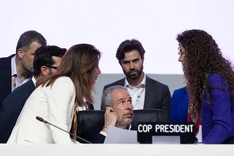 Un grupo de personas, entre ellas un hombre sentado detrás de un escritorio con la etiqueta COP PRESIDENT, participan en un debate sobre combustibles fósiles en una conferencia o reunión oficial. El fondo es claro y claro.