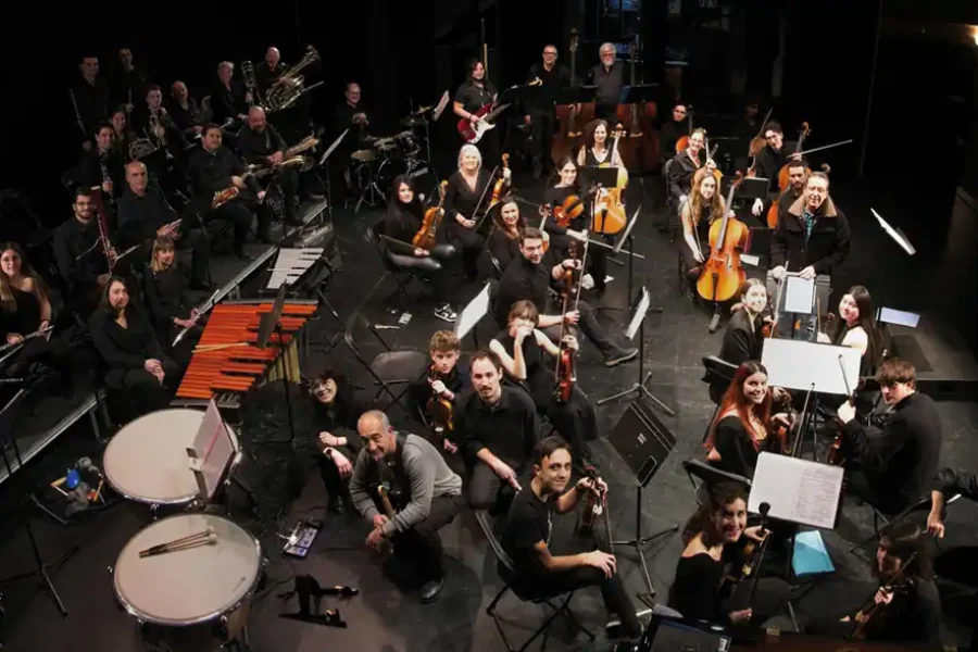 La Orquesta Athanor está sentada y de pie en un escenario oscuro de Rivas, con cuerdas, metales y percusión. Los músicos están colocados por secciones, de cara a la cámara y mirando hacia arriba, listos para interpretar el Carnaval de los animales.