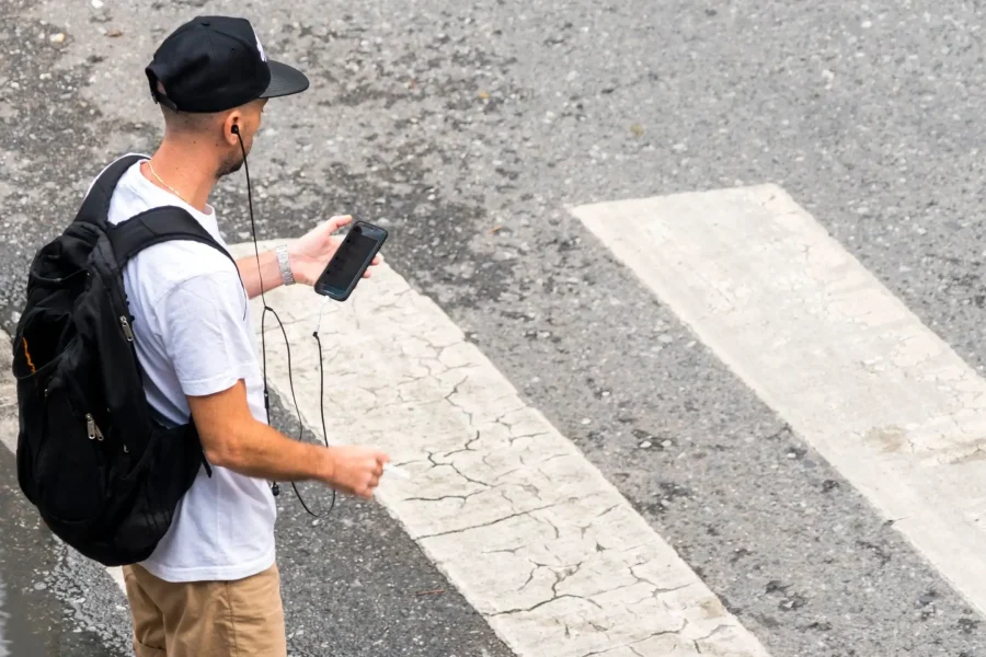 Una persona con gorra negra, camiseta blanca, pantalones cortos de color canela y mochila se para en un paso de peatones con un teléfono en la mano y auriculares en los oídos, mirando hacia la carretera, dispuesta a cruzar calles donde el pavimento desgastado marca la guía de los peatones.