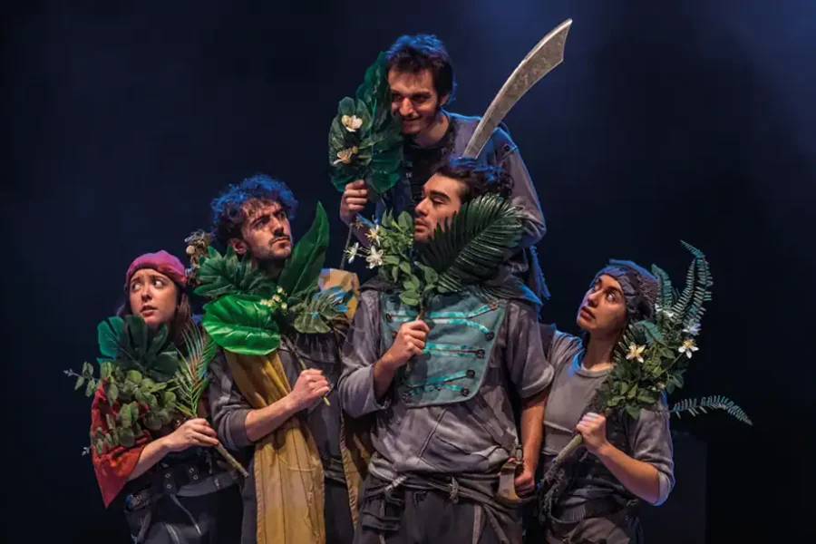 Durante el estreno en Rivas, cinco actores disfrazados se sitúan en el escenario sosteniendo grandes hojas verdes y plantas. Un actor, elevado sobre los demás, empuña una gran espada mientras todos adoptan poses expresivas y miran en distintas direcciones.