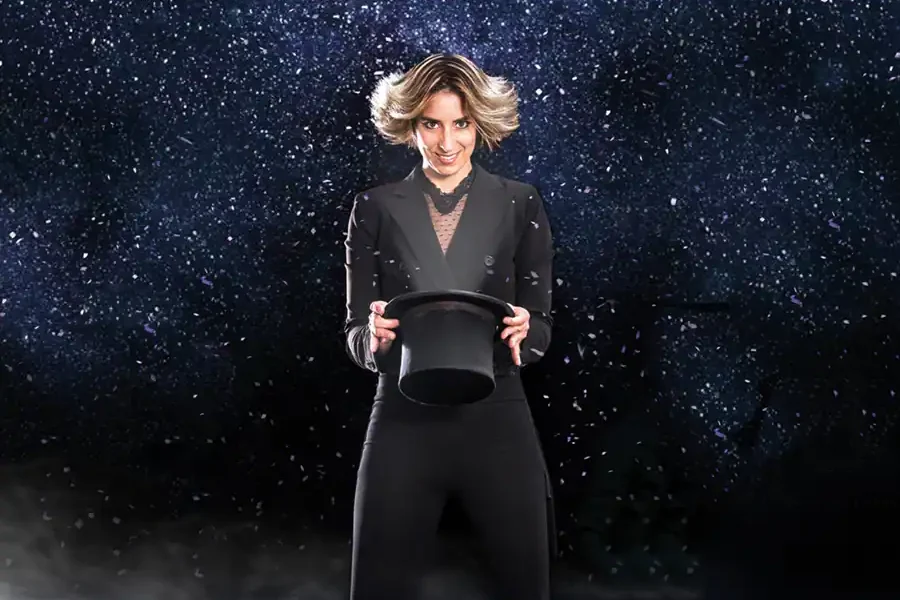 En el Festival de Magia de Rivas, una persona vestida de negro, sobre un fondo oscuro y estrellado, sostiene un sombrero de copa negro mientras partículas de nieve o confeti flotan en el aire, evocando la magia de la Navidad.