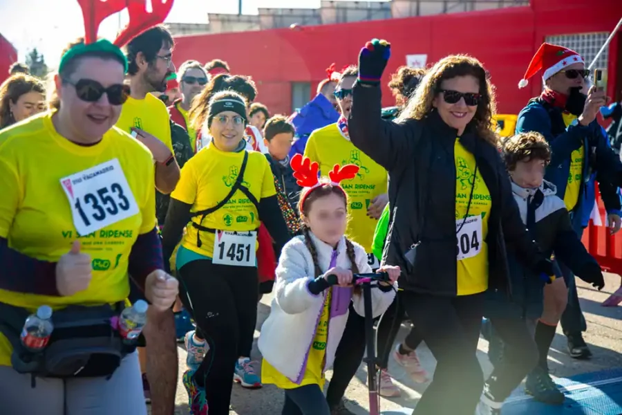Un grupo de personas con camisetas amarillas y dorsales numerados participan en la carrera popular San Ripense en Rivas. Una mujer levanta el puño, mientras un niño con cuernos rojos de reno monta en patinete. Otros sonríen y caminan juntos.