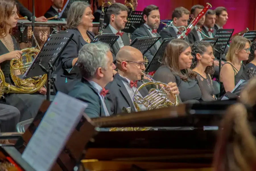 Músicos en traje de etiqueta actúan en la orquesta La Sinfónica de Rivas, tocando instrumentos de viento-metal y madera. Se ven atriles con partituras y, al fondo, más músicos e instrumentos en el Abono Sinfónico 2026.