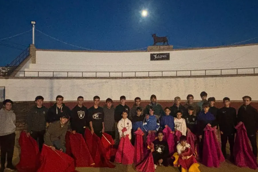 Un grupo de unas 25 personas, en su mayoría niños y adolescentes, posan juntos en una plaza de toros al aire libre al anochecer, cada uno con un capote rojo o rosa. Una luna y una estatua de un toro se ven en el borde superior del muro de la plaza.