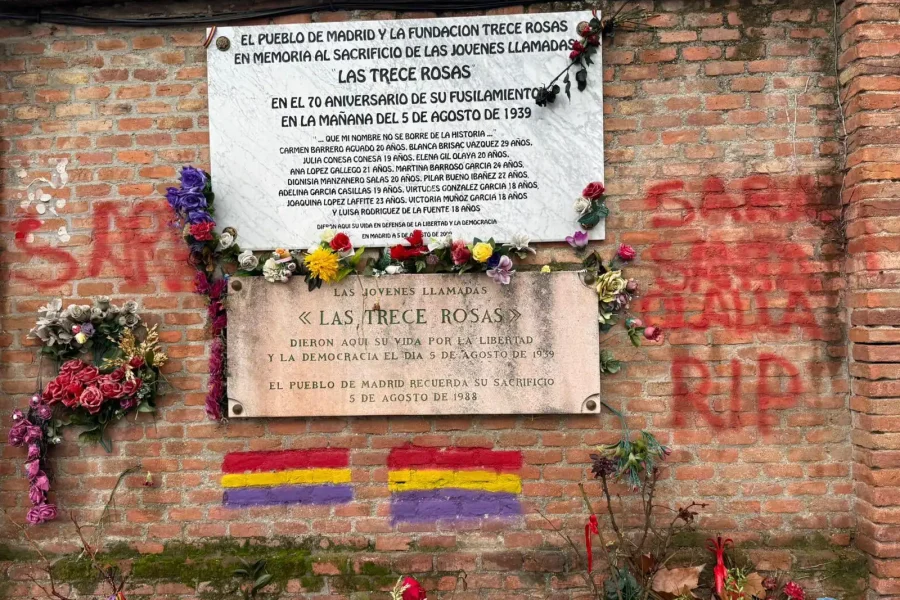 Un muro de ladrillo muestra dos placas y flores conmemorativas de Las Trece Rosas. Se ven pancartas, pintadas y amenazas rojas. Ambas placas -escritas en español- honran a las jóvenes ejecutadas en 1939 en la tumba de las 13 Rosas.