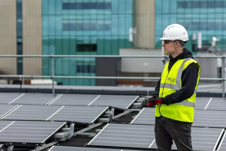 Una persona con casco blanco, gafas de seguridad y chaleco amarillo de alta visibilidad se encuentra entre paneles solares en un tejado, comprobando un borrador automático, con un moderno edificio al fondo.