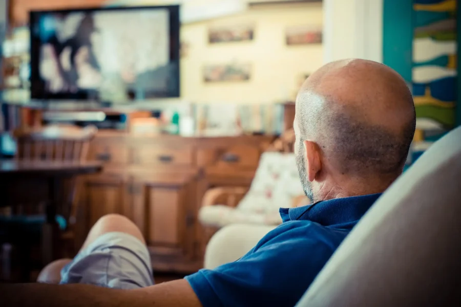 Un hombre calvo con camisa azul está sentado en el sofá de su salón, viendo la televisión, tal vez perdido en atracones de serie. El televisor aparece difuminado en el fondo, junto con muebles de madera y objetos decorativos.