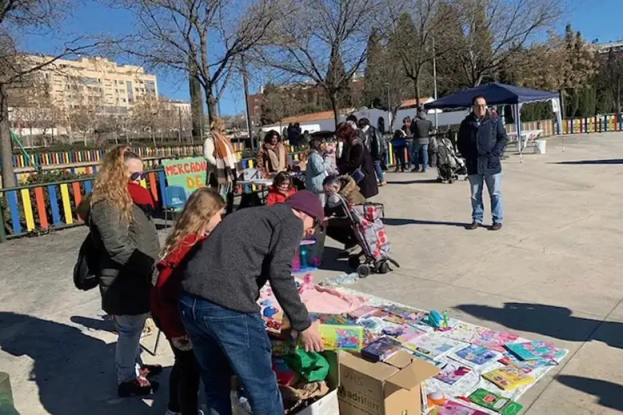 La gente se reúne al aire libre en la soleada Rivas, mirando los artículos expuestos para la venta o el intercambio. Algunos hacen cola, mientras otros interactúan con los productos. Al fondo se ven una valla de colores y varios edificios que contribuyen a crear un ambiente animado.