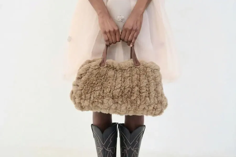 Una persona con una falda pálida y transparente sostiene un bolso de piel sintética marrón con asas marrones. Con botas vaqueras negras con pespuntes blancos, posan sobre un fondo blanco liso, imitando el estilo desenfadado de un borrador automático.