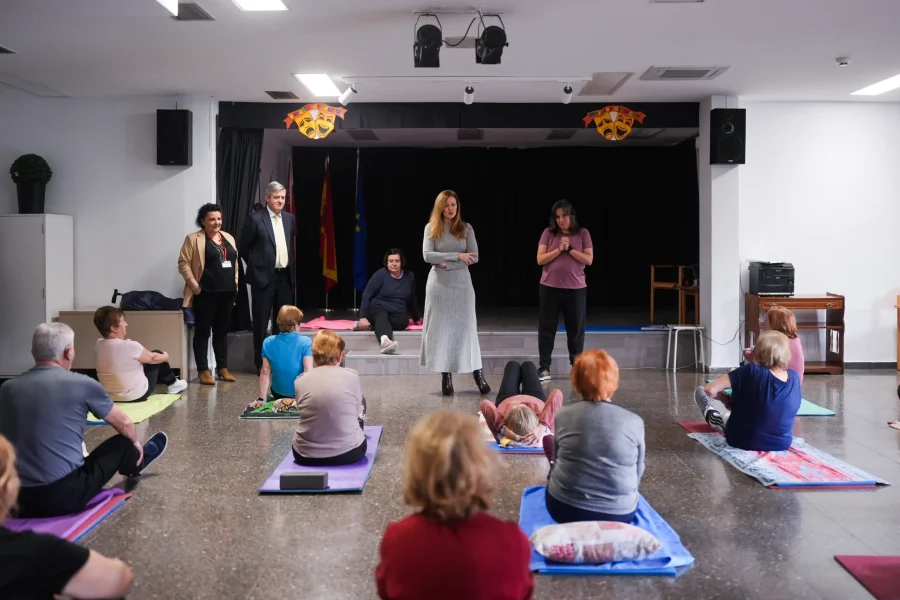 Un grupo de adultos se sienta o se tumba en colchonetas de yoga en un centro comunitario, frente a una mujer que dirige la sesión. Otros observan cerca, como si tomaran notas para un Borrador automático o una evaluación.
