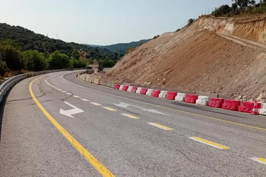 Una carretera de dos carriles traza una curva a la izquierda con claras marcas de carril amarillas y blancas, que recuerdan a un borrador automático. Barreras rojas y blancas bordean el lado derecho cerca de una ladera con suelo expuesto, mientras que al fondo se ven árboles y colinas.