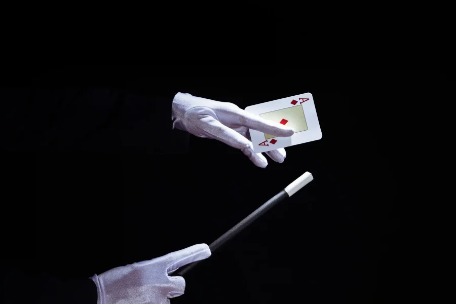 Una persona con guantes blancos sostiene dos naipes, el as de diamantes y el as de corazones, en una mano y con la otra apunta a los naipes con una varita de mago negra, captando un momento de magia en Rivas sobre un fondo negro.
