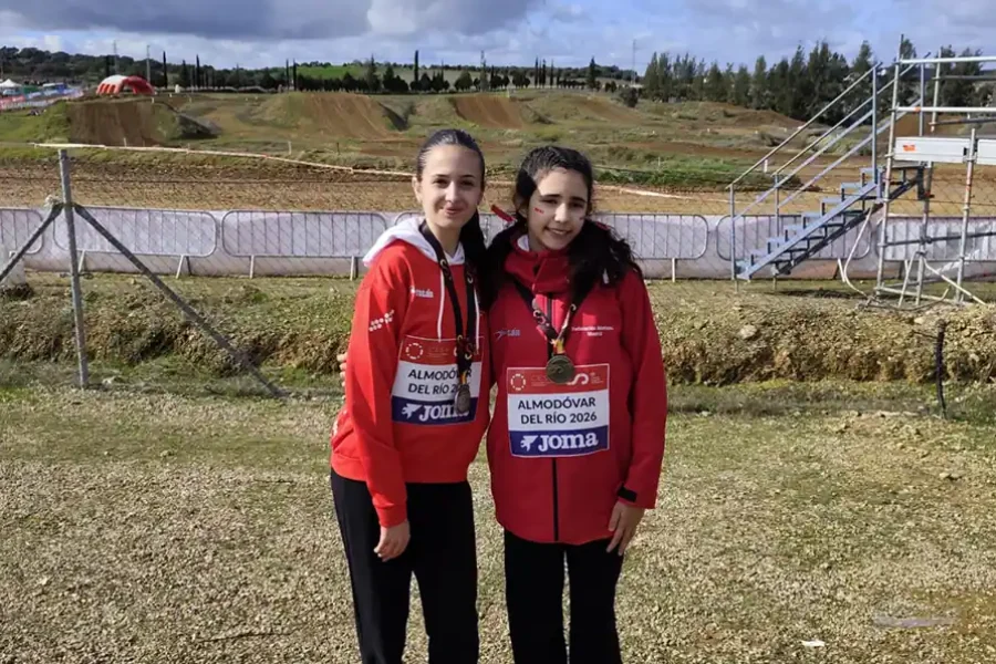 Dos jóvenes atletas con chaquetas rojas y pantalones negros posan uno al lado del otro en un cross estatal en Ciudad de Rivas, posando en un campo de hierba con dorsales, barreras metálicas y escaleras visibles al fondo.