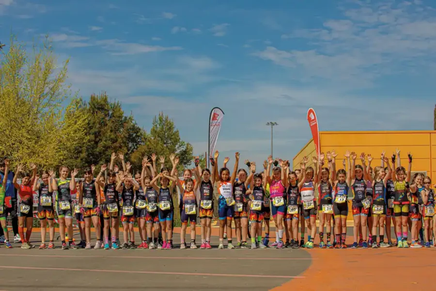 Un grupo de niños con ropa deportiva forman una fila al aire libre con los brazos en alto, preparados para el Duatlón Escolar de Rivas. Hay árboles, banderas y un edificio amarillo de fondo bajo un cielo azul mientras esperan sus categorías y fechas.