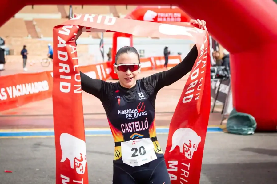 Una triatleta con gafas de sol y dorsal 20 sostiene una pancarta roja en la línea de meta en la que se lee TRIATLON MADRID, celebrando su victoria mientras el borrador automático capta el momento triunfal.
