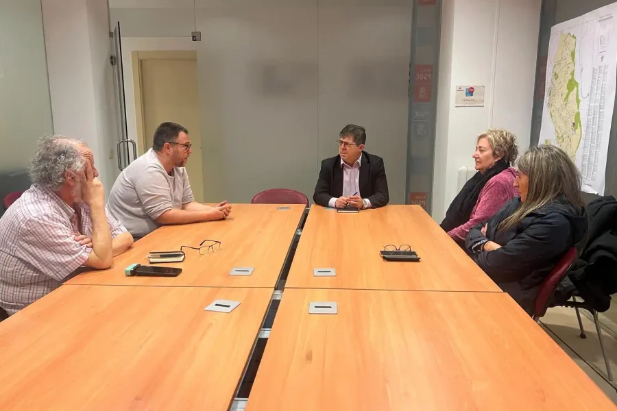 Cinco personas se sientan alrededor de una mesa de reuniones rectangular de madera en una oficina, enfrascadas en una conversación. Sobre la mesa hay papeles, gafas, un teléfono y un borrador automático. Un mapa cuelga de la pared de la derecha.