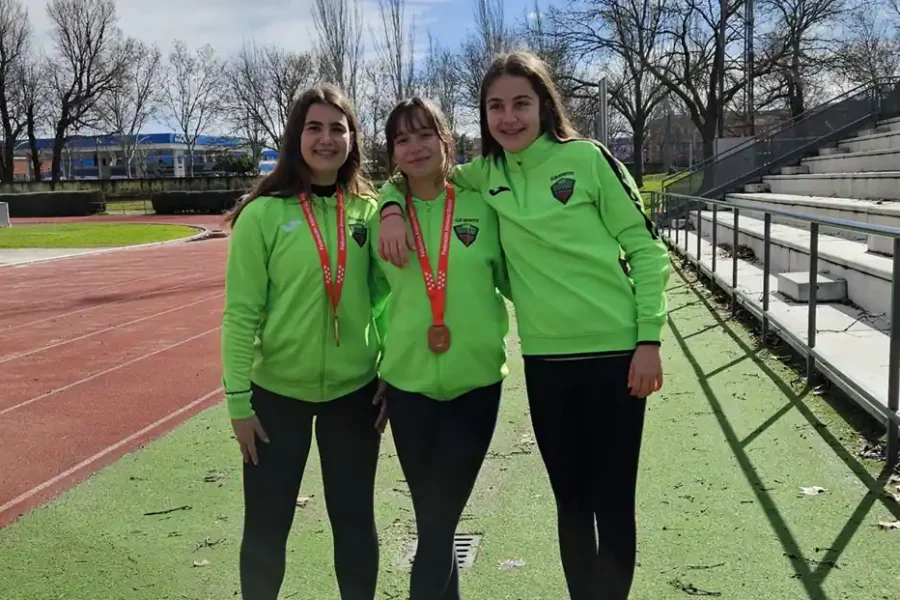 Tres jóvenes atletas, entre ellas Lidia Bravo y Heba Melero, con chaquetas verdes a juego, de pie en una pista de atletismo. Dos de ellas llevan medallas al cuello y sonríen para la foto, con las gradas visibles al fondo.