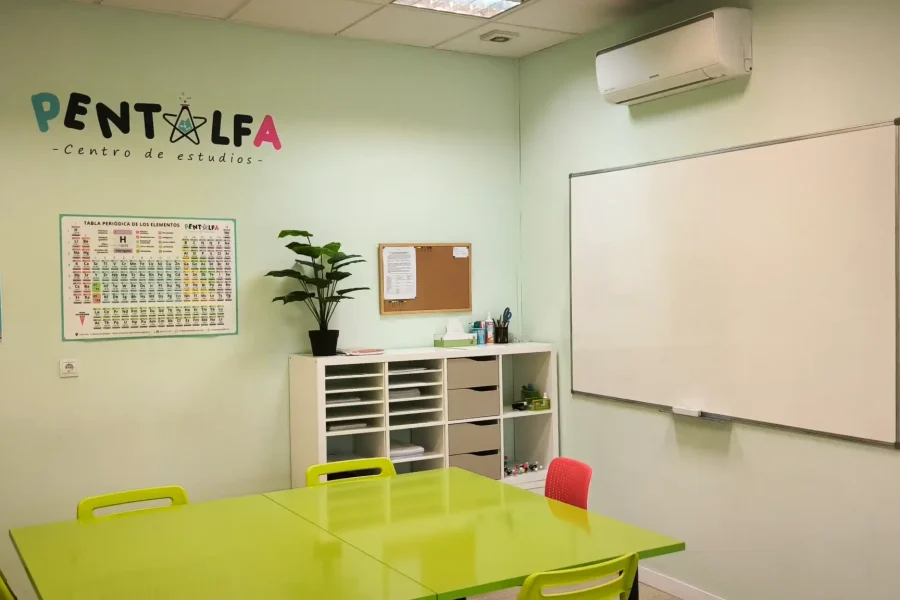 Un aula de la academia Pentalfa tiene paredes verde pálido, una mesa verde lima con sillas, una pizarra blanca, una tabla periódica, un mapamundi, una planta, estanterías con material y un tablón de anuncios, ideal para el aprendizaje escolar. "PENTALFA" está pintado en la pared.