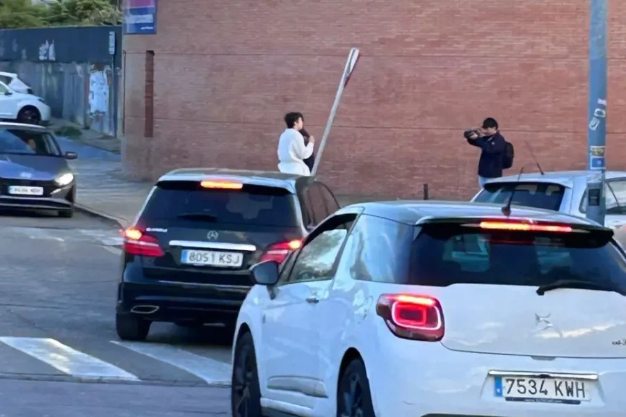 Un hombre con camisa blanca se encuentra en la acera sosteniendo una señal de tráfico cerca de un mural de sanidad pública, mientras otra persona le hace una foto. Varios coches esperan en un paso de peatones frente al edificio de ladrillo.