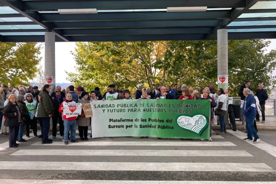 Un grupo de personas en un paso de peatones con una pancarta que dice Sanidad pública de calidad es dignidad y futuro para nuestros pueblos en una manifestación pública, exigiendo soluciones a la falta de personal en el Hospital del Sureste, con árboles cerca.