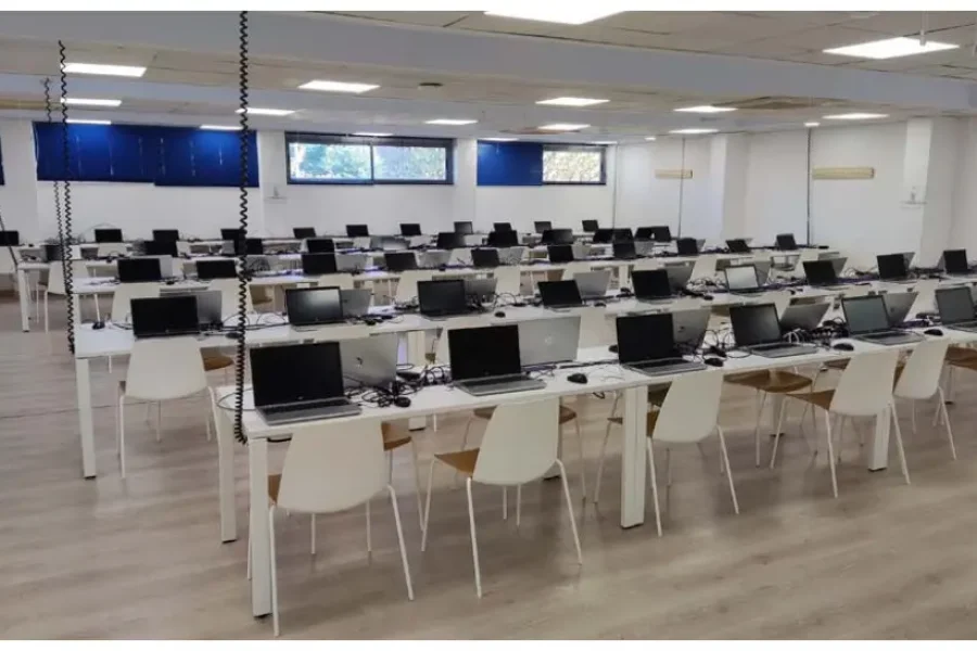Una amplia sala con filas de mesas y sillas blancas, cada puesto equipado con un ordenador portátil, un teclado, un ratón y un monitor. Ideal para los eventos de la bolsa de empleo de Rivamadrid, la sala cuenta con persianas azules y una iluminación cenital muy luminosa.