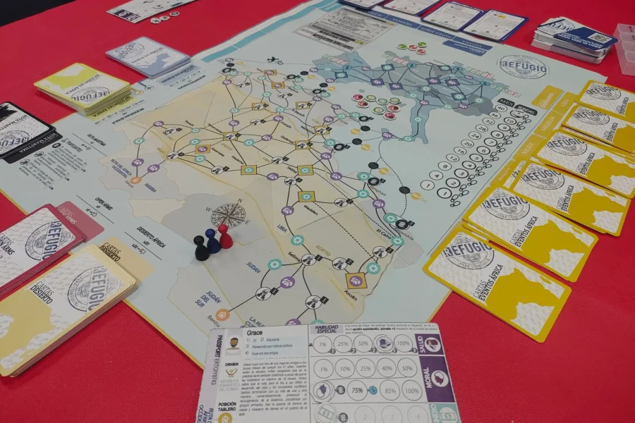 En una mesa roja de Ludiversia hay un juego de mesa con un mapa con rutas, fichas de colores, varios mazos de cartas y cartas de jugador de personaje. Varias fichas invitan a probar juegos con amigos.