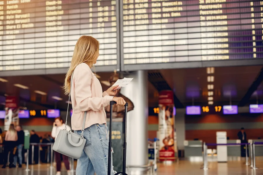 Una mujer se encuentra en un aeropuerto de España, con una tarjeta de embarque y una maleta en la mano, mirando un gran panel de salidas en el que se muestra la información de los vuelos para los pasajeros aéreos internacionales. Lleva ropa informal y un bolso ligero, preparada para su viaje de este mes de julio.