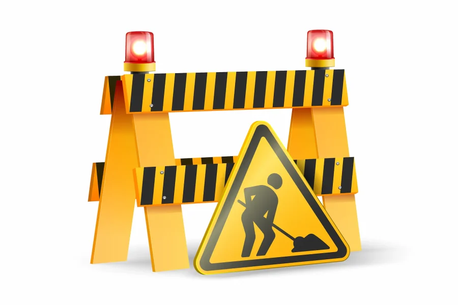 Una barrera amarilla de construcción con rayas negras y dos luces rojas de advertencia se encuentra cerca de una señal amarilla triangular que muestra las obras de carretera por delante, la señalización de obras en la rotonda Pilar Miró, según informa la Asociación Vecinal Ladera del Almendro.