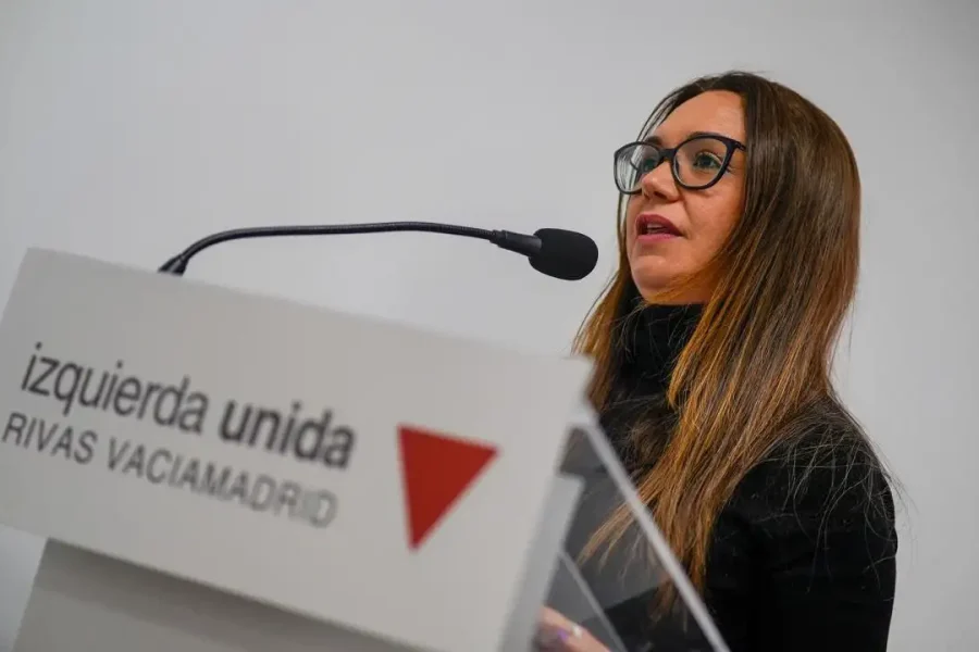 Aída Castillejo, coordinadora de IU Rivas, con pelo largo castaño y gafas, habla en un estrado rotulado izquierda unida RIVAS VACIAMADRID con un logotipo de un triángulo rojo, dirigiéndose al micrófono sobre un fondo liso y claro.