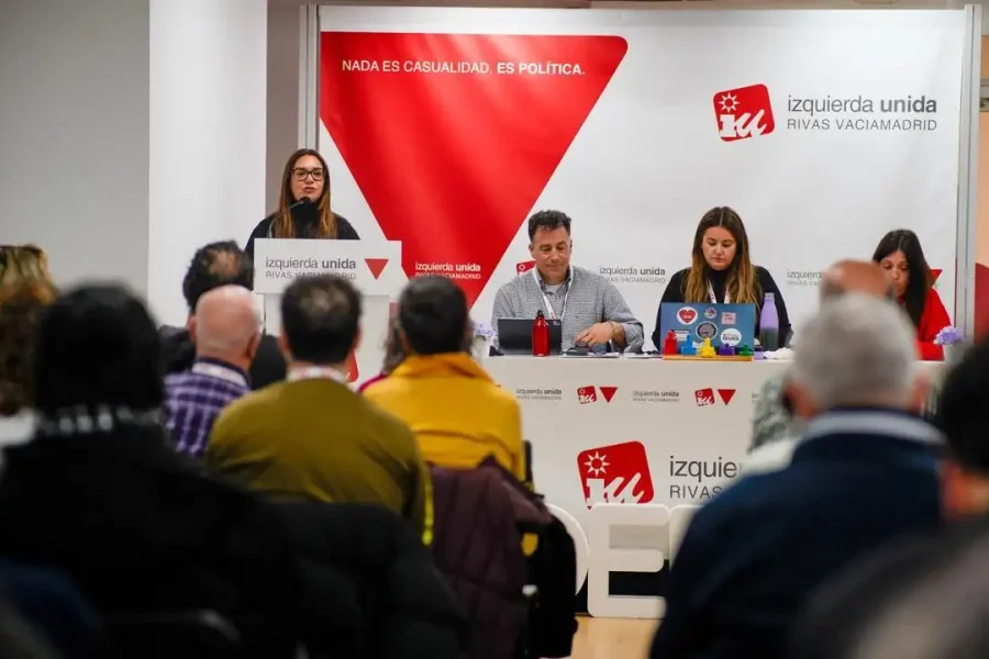 Aída Castillejo, coordinadora de IU Rivas, habla en un podio a un público en una sala de conferencias, con tres personas sentadas en una mesa a su lado. Por toda la sala se ven pancartas y carteles con "izquierda unida" y gráficos rojos.