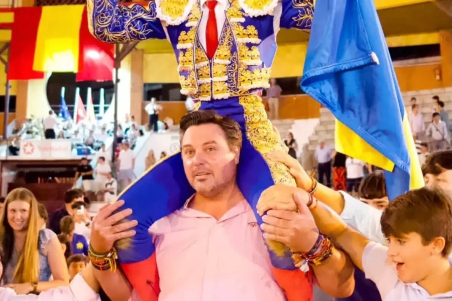 En el Circuito de Madrid 2026, una persona vestida con un traje ornamentado azul y dorado es levantada a hombros por alguien, mientras sostiene un trofeo y una bandera azul y amarilla; los espectadores vitorean a Sergio Rollón entre coloridas pancartas.