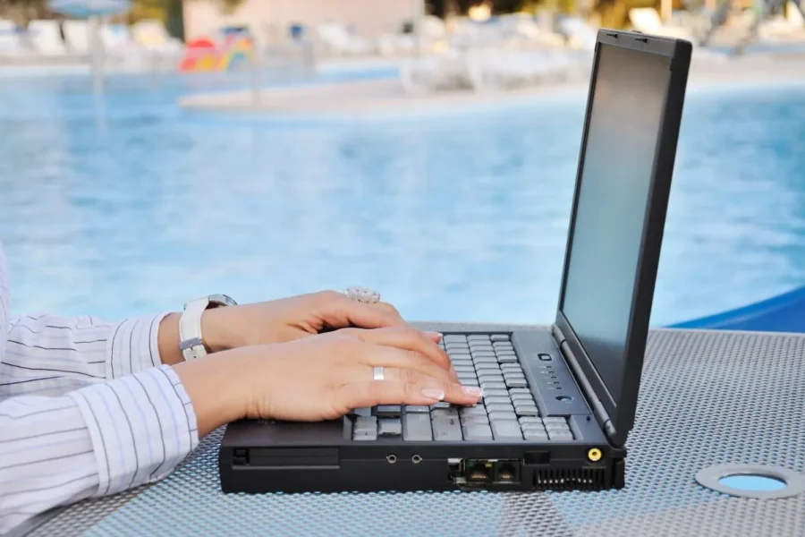 Una persona teclea en un portátil colocado sobre una mesa metálica junto a una piscina, reflejando cómo los españoles buscan desconectar del trabajo en verano. El fondo muestra agua cristalina, sillas junto a la piscina y un entorno exterior difuminado.