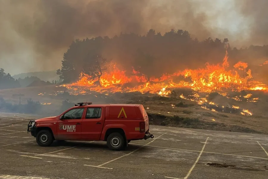 Un vehículo rojo de respuesta a emergencias está aparcado en un terreno vacío mientras las grandes llamas y el denso humo de los incendios arden entre la maleza y los árboles del fondo.