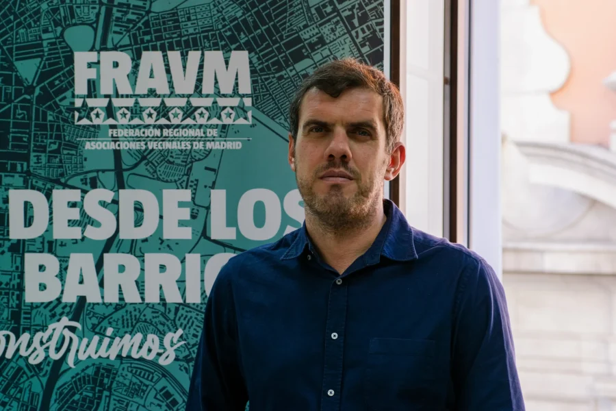 Jorge Nacarino, un hombre de pelo corto y castaño, con camisa azul, permanece de pie en un interior, delante de una pancarta verde azulado del Fravm con texto en blanco en español y un mapa gráfico de Madrid. La luz del sol entra por una ventana a la derecha, dejando entrever el vibrante ruido político de la ciudad.