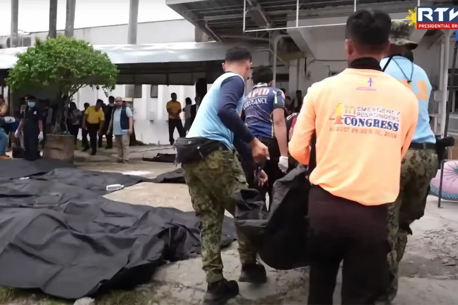 Varios equipos de emergencia y agentes de policía manipulan bolsas negras para cadáveres al aire libre cerca de un edificio en Filipinas, tras un terremoto, mientras los espectadores permanecen de pie al fondo; se ven lonas o más bolsas para cadáveres en el suelo.