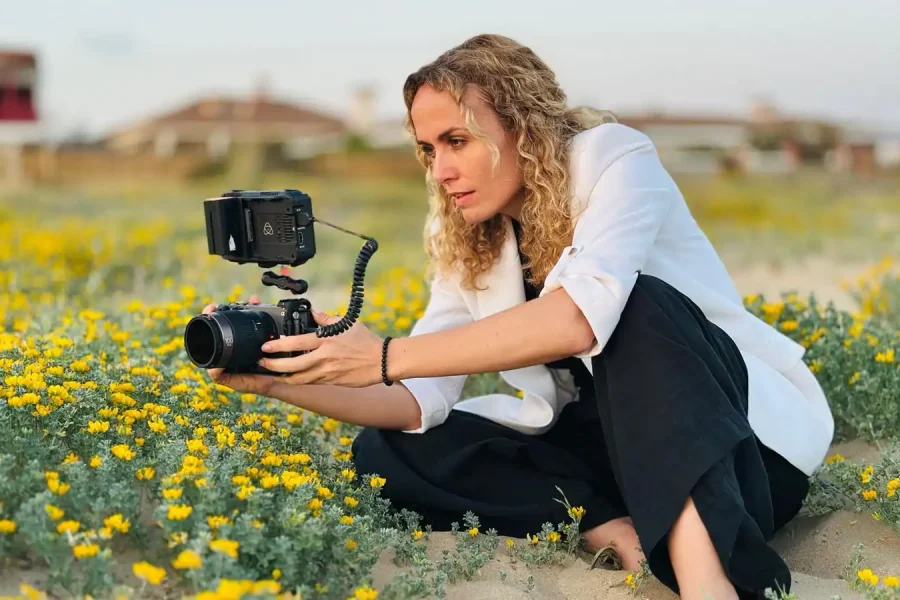 Una persona con el pelo rizado, vestida con una americana blanca y pantalones negros, se sienta entre flores silvestres amarillas mientras sostiene una cámara con un monitor conectado, captando la escena, tal vez contando historias de víctimas o destacando la desprotección judicial.
