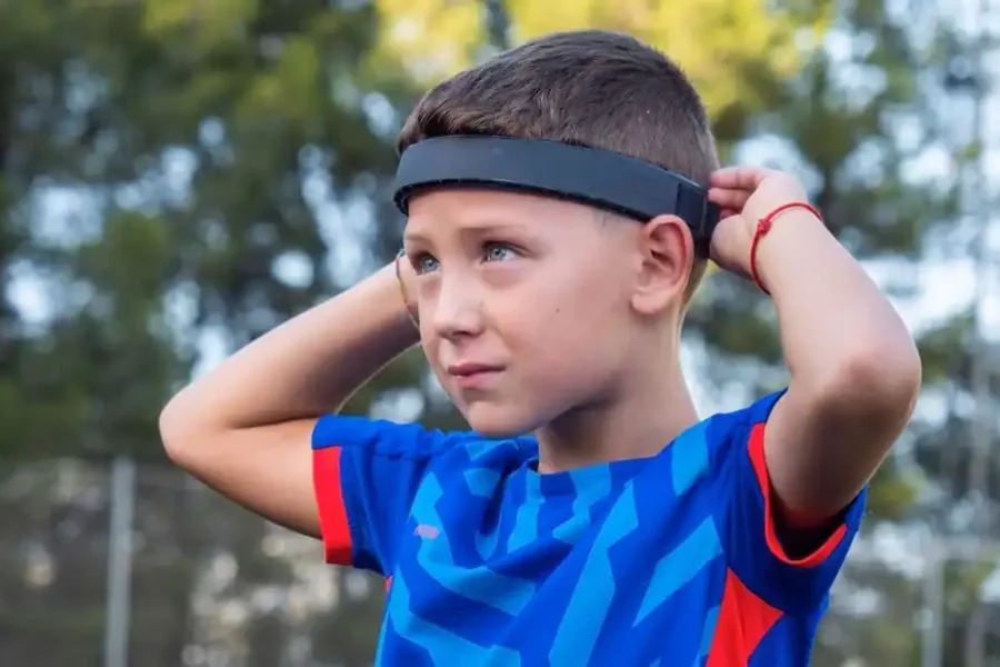 Un niño con una camiseta deportiva azul se ajusta una cinta negra en la cabeza al aire libre, con árboles y una valla en el fondo borroso, destacando cómo los niños futbolistas se protegen durante los remates de cabeza para ayudar a prevenir posibles daños neurológicos.