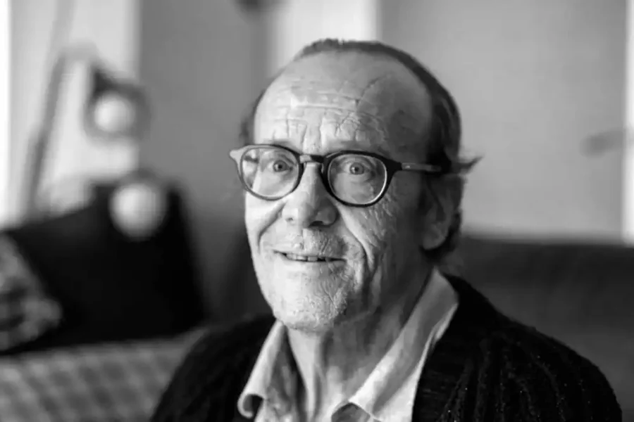 Un hombre mayor con gafas y el pelo ralo está sentado en el interior, mirando a la cámara y sonriendo ligeramente. La foto, en blanco y negro, tiene un fondo borroso con una lámpara y un cojín que evocan historias de la Puerta del Sol durante la época del expreso del franquismo.