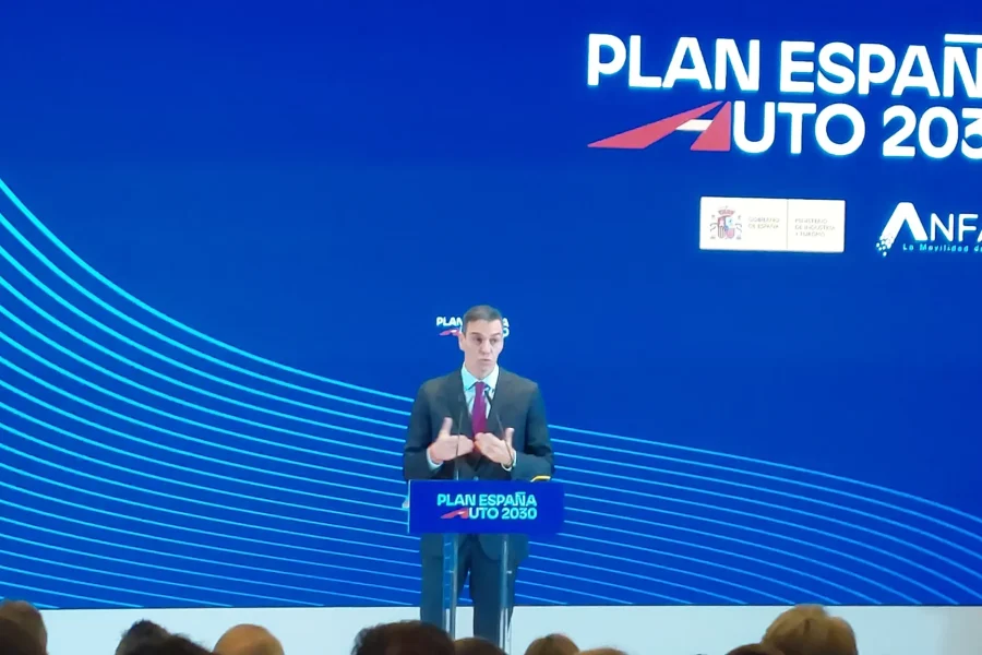 Una persona, de pie en un podio con el rótulo Plan España Auto 2030, se dirige al público para hablar de las ayudas directas y el despliegue de recargas. Detrás, una gran pantalla azul muestra el Plan España Auto 2030, logotipos de ANFAC y referencias a iniciativas relacionadas con el coche eléctrico.