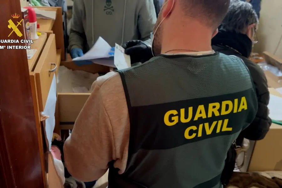Un agente de la Guardia Civil ataviado con un chaleco en el que se lee Guardia Civil permanece de pie frente a un cajón abierto mientras otra persona manipula documentos en una sala durante una inspección relacionada con una red criminal de estafas online.