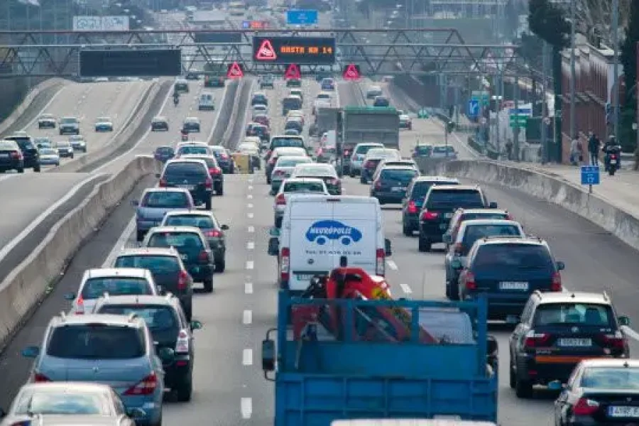 Varios carriles de coches y camiones soportan un intenso tráfico durante la operación salida en una autopista. Señales electrónicas en lo alto muestran mensajes, mientras que barreras de hormigón separan los sentidos de circulación. Edificios y árboles se alinean a los lados de la carretera.