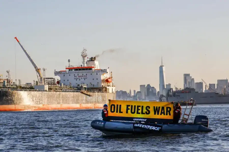 Un pequeño barco de Greenpeace con tres personas sostiene una gran pancarta amarilla en la que se lee OIL FUELS WAR en el agua cerca de una gran nave industrial, destacando cómo los combustibles fósiles impulsan la espiral climática, con el horizonte de una ciudad al fondo.