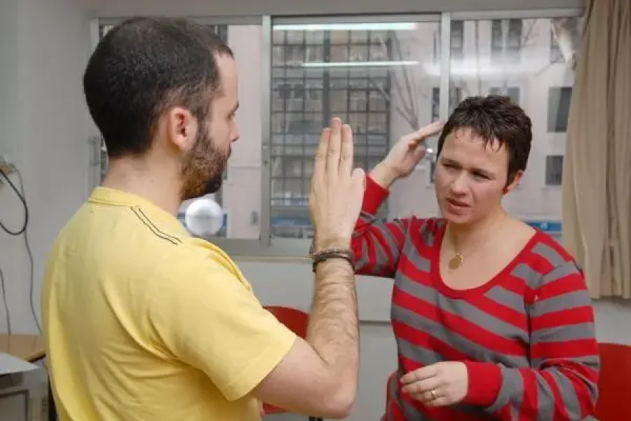 Dos personas se encuentran frente a frente en un interior. La persona de la izquierda levanta una mano, mientras que la de la derecha, con camisa roja a rayas, hace un gesto cerca de la cabeza, posiblemente utilizando Lengua de Signos Española. Al fondo se ven una ventana y sillas.