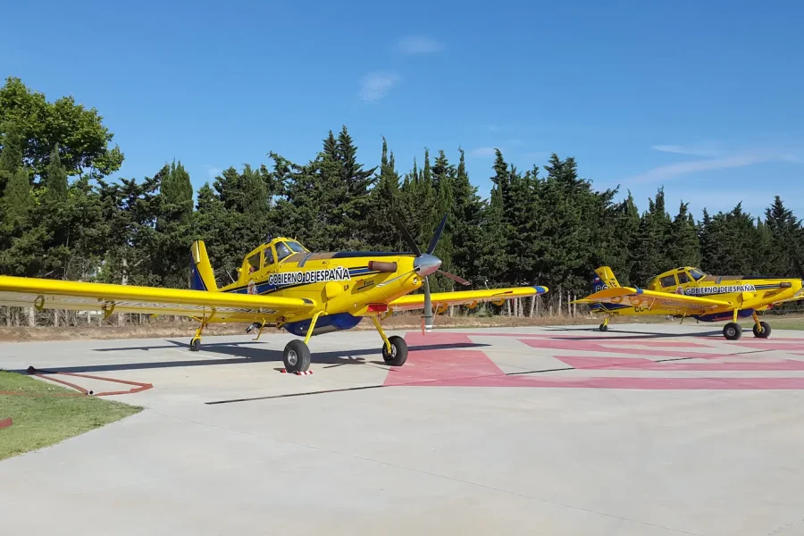 Dos aviones monomotores de color amarillo brillante, con la marca BOMBEROS DE SEGORBE, están estacionados sobre una superficie de hormigón con marcas rojas. Rodeados de árboles verdes y un cielo azul despejado, estos aviones cuentan con un sistema de borrador automático para mayor eficacia.