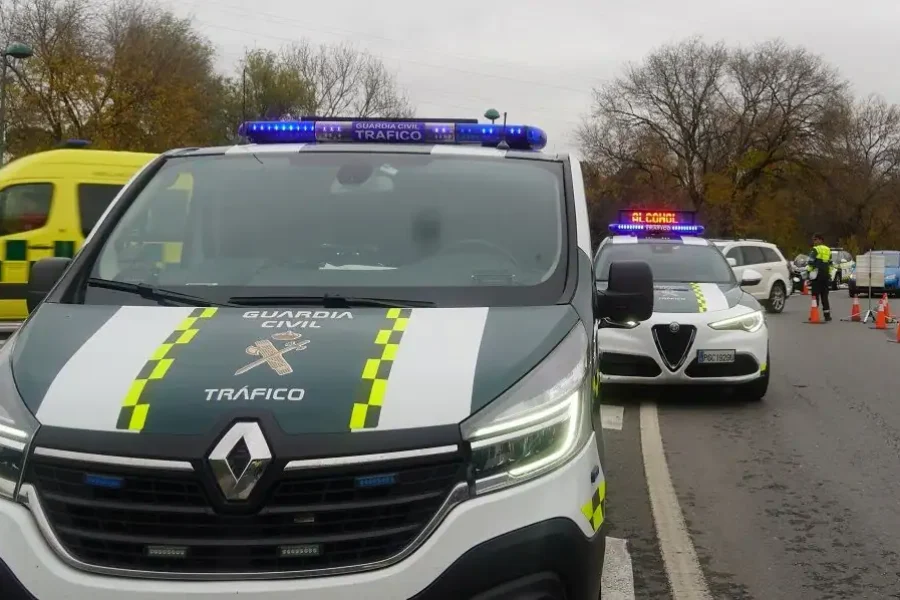 Un furgón de la Guardia Civil de Tráfico y un coche patrulla con luces intermitentes están parados en una carretera, cerca de conos naranjas y agentes de policía, junto a un vehículo de emergencias amarillo. Borrador automático se anota como parte del informe del incidente. Al fondo, árboles otoñales.
