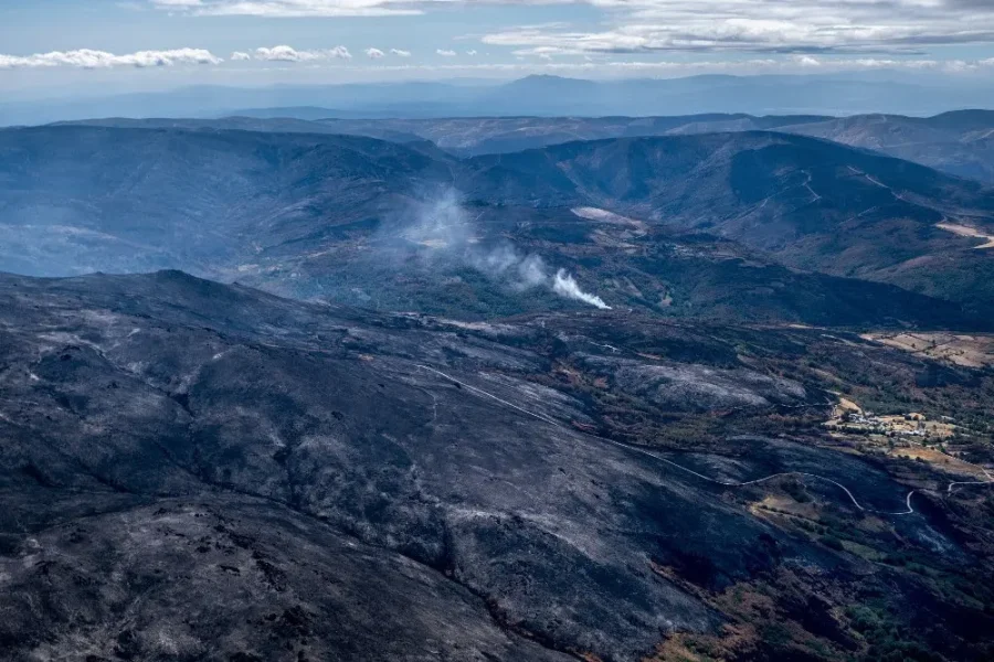 Vista aérea de un paisaje montañoso con grandes extensiones de tierra calcinada y ennegrecida y manchas de humo que se elevan desde el bosque, lo que indica los daños provocados por un reciente incendio forestal. En este borrador automático se aprecia algo de vegetación verde y un pequeño asentamiento.