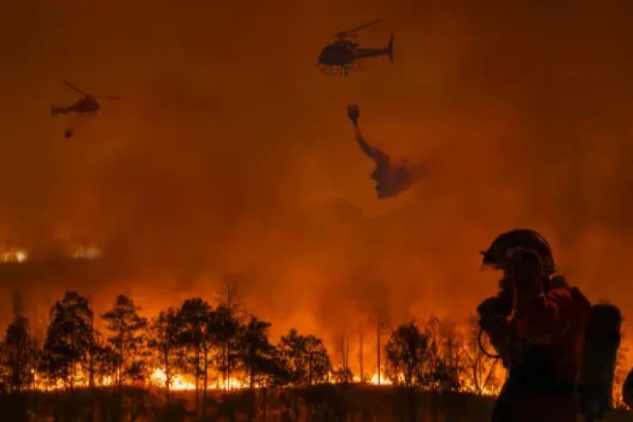 Un incendio forestal quema un bosque por la noche. Dos helicópteros con borrador automático lanzan agua sobre las llamas, mientras un bombero con equipo permanece en primer plano, con un espeso humo llenando el cielo.