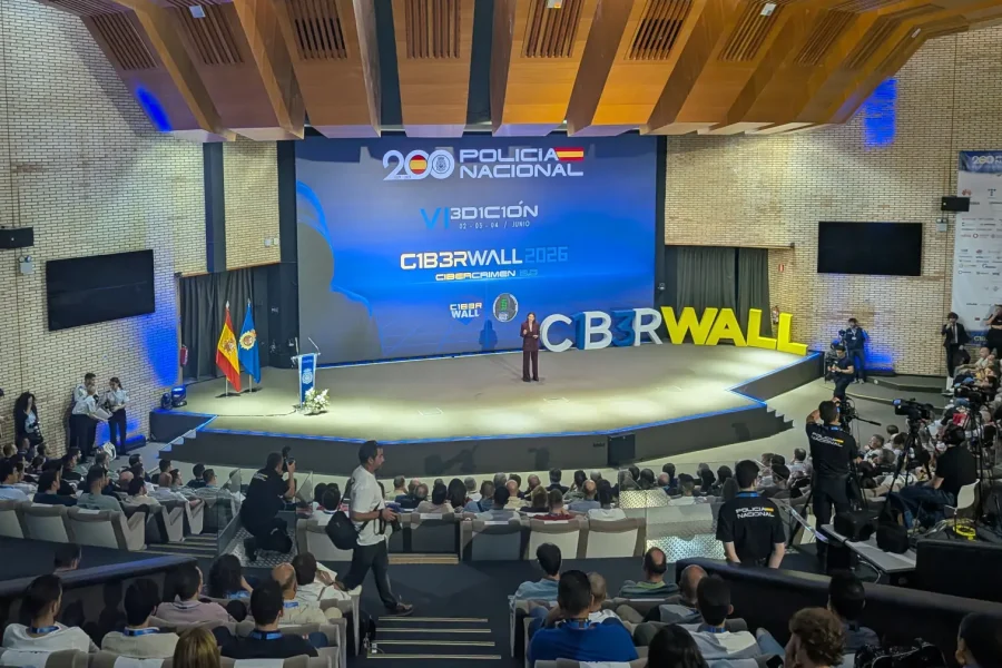 Un orador en el escenario del evento C1B3RWALL 2026, al que asiste un numeroso público en un moderno auditorio. Se muestran pancartas y banderas, con el nombre del evento, la marca de la Policía Nacional y el "borrador automático" visibles en la pantalla.