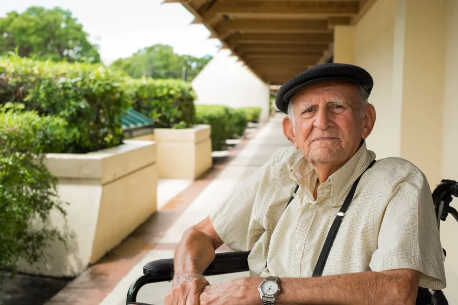 Un anciano que lleva una gorra plana, una camisa de manga corta a rayas y un reloj de pulsera está sentado en una silla de ruedas junto a una acera, lo que pone de manifiesto la importancia del apoyo para las personas con dependencia, con vegetación y una acera cubierta al fondo.
